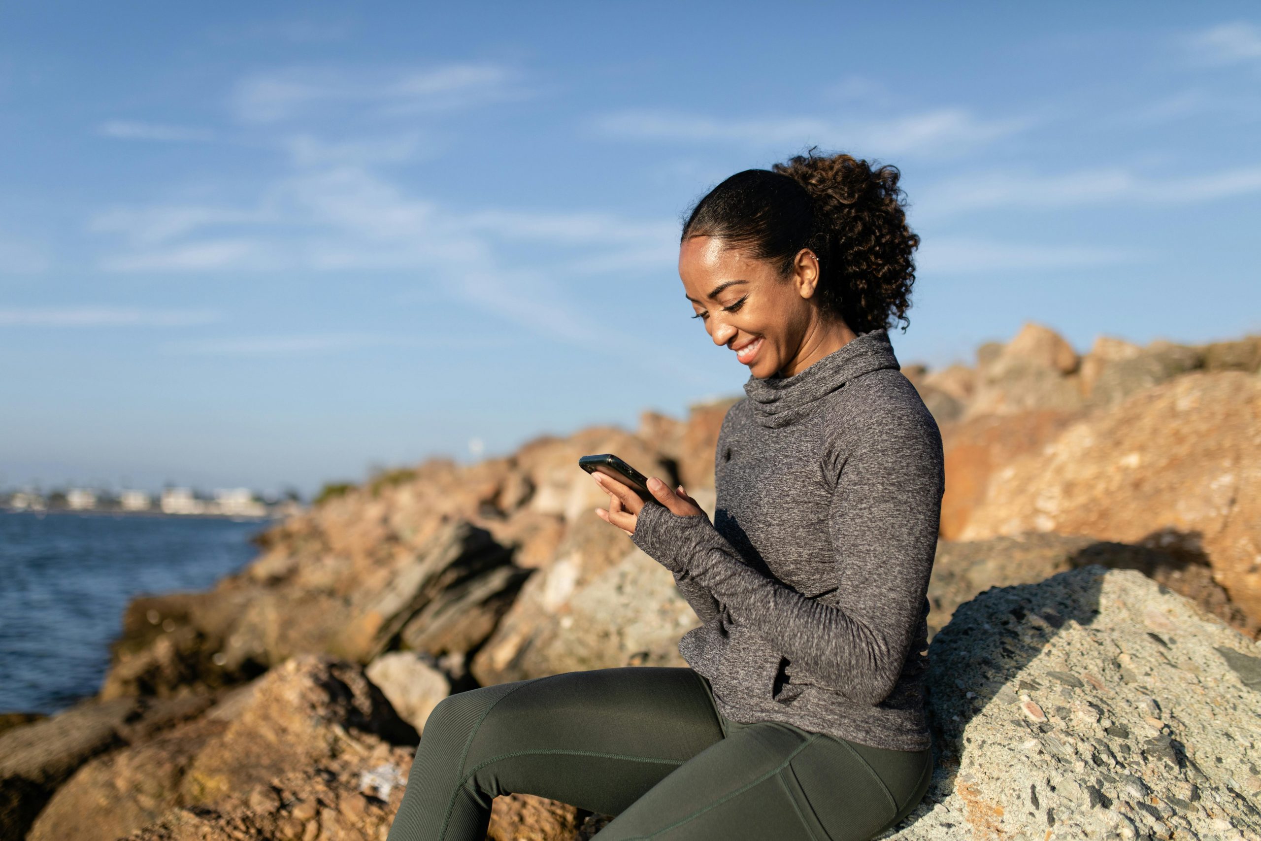 Happy woman sitting on rocks by the ocean, using her phone to share her experience